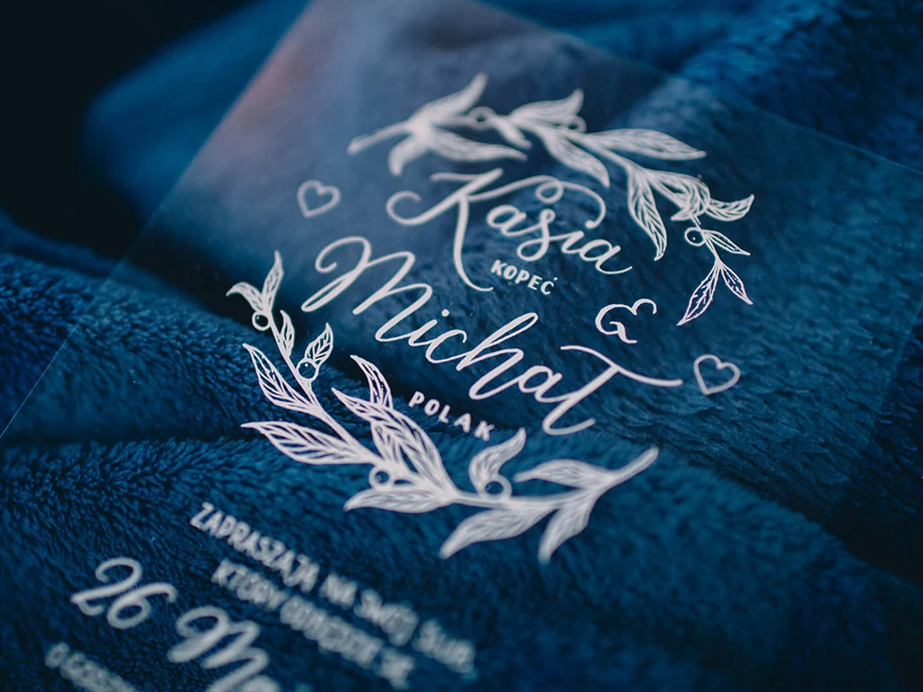 A transparent wedding invitation printed in white hand lettering with a botanical wreath, photographed on dark blue fabric.